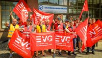 Gruppe von etwa 15 Personen steht dicht beieinander vor einem Gebäude mit Backsteinfassade und großen Fenstern. Mehrere Personen tragen orangefarbene oder gelbe Warnwesten. In den Händen werden rote Fahnen und Banner gehalten, auf denen in weißen Buchstaben „EVG“ sowie „Eisenbahn- und Verkehrsgewerkschaft“ steht. Einige Personen heben die Arme. Hinter der Gruppe ist ein Schild mit der Aufschrift „GLEISBAUMECHANIK“ sowie eine Adresse und Telefonnummer sichtbar.