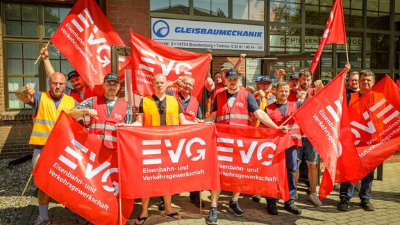 Gruppe von etwa 15 Personen steht dicht beieinander vor einem Gebäude mit Backsteinfassade und großen Fenstern. Mehrere Personen tragen orangefarbene oder gelbe Warnwesten. In den Händen werden rote Fahnen und Banner gehalten, auf denen in weißen Buchstaben „EVG“ sowie „Eisenbahn- und Verkehrsgewerkschaft“ steht. Einige Personen heben die Arme. Hinter der Gruppe ist ein Schild mit der Aufschrift „GLEISBAUMECHANIK“ sowie eine Adresse und Telefonnummer sichtbar.