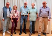 Fünf Personen stehen in einer Reihe vor einem weißen Vorhang in einem Innenraum mit Holzfußboden. Die Personen tragen überwiegend Freizeitkleidung. Im Hintergrund sind Vorhänge und links ein Teil eines Heizkörpers zu sehen.