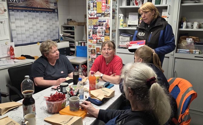 Eine Gruppe von Menschen sitzt um einen Tisch in einem Aufenthaltsraum. Auf dem Tisch stehen Getränke, Tassen, Snacks und mehrere kleine Papiertüten. Eine weitere Person steht am Tischrand und hält eine Packung mit Gebäck oder Süßwaren. Im Hintergrund sind Regale, ein großer Wandkalender und verschiedene Aushänge sowie Gegenstände des Raums zu sehen.