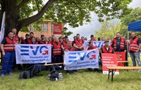 Gruppe von etwa 25 Personen steht im Freien auf einer Wiese unter einem großen Baum. Viele tragen rote Warnwesten mit dem Logo „EVG“. Mehrere Personen halten weiße Banner mit blau-rotem Logo und der Aufschrift „EVG Eisenbahn- und Verkehrsgewerkschaft“. Im Hintergrund sind rote Plakate zu sehen mit dem Text „Heute ist kein Arbeitstag. Heute ist STREIKTAG!“. Hinter der Gruppe steht ein Backsteingebäude mit Fenstern. Im Vordergrund liegen Taschen und Jacken auf dem Boden, daneben stehen Bierbänke und -tische.