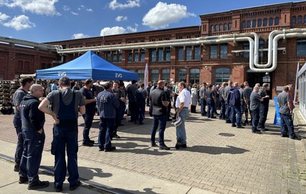 Gruppe von Personen auf einem gepflasterten Hof vor einem großen Backsteingebäude. Ein blaues Zelt mit der Aufschrift „EVG“ steht im linken Bildbereich. Die Personen stehen in kleinen Gruppen zusammen. Über dem Gebäude ist ein blauer Himmel mit einzelnen Wolken zu sehen.
