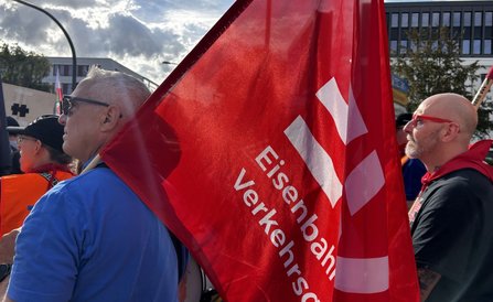 Nahaufnahme einer roten Fahne mit weißem Schriftzug „Eisenbahn- und Verkehrs…“ im Vordergrund. Daneben stehen mehrere Personen, teilweise mit orangefarbenen Warnwesten. Im Hintergrund sind Gebäude, Straßenschilder und ein bewölkter Himmel mit Sonnenlicht zu sehen.