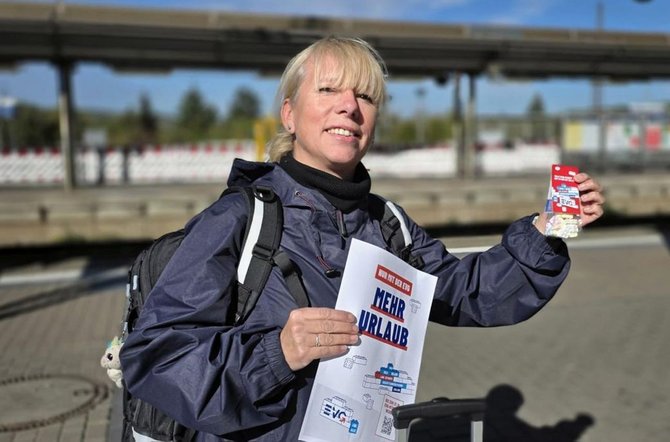 Person mit Rucksack steht auf einem Bahnsteig und hält einen Flyer mit der Aufschrift „NUR MIT DER EVG MEHR URLAUB“ sowie eine kleine rote Verpackung in den Händen. Im Hintergrund sind Bahnsteiganlagen und Gleise zu sehen.
