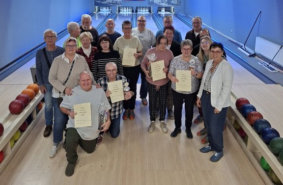 Gruppenfoto auf einer Kegel- oder Bowlingbahn: 17 Personen stehen und knien auf der Bahn zwischen zwei Kugelrückläufen mit mehreren farbigen Kugeln. Mehrere Personen halten Urkunden, einige zusätzlich kleine Pokale oder eingepackte Präsente. Im Hintergrund sind mehrere Bahnen mit aufgestellten Kegeln sichtbar. Die Halle ist hell beleuchtet, mit blauen Lichtstreifen entlang der Bahnen und hellen Wänden.