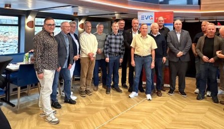 Gruppenfoto in einem Innenraum mit Parkettboden. Etwa 13 Personen stehen in zwei Reihen vor einem Roll-up-Banner mit dem sichtbar lesbaren Schriftzug „EVG“. Im Hintergrund sind Fenster, Tische und Stühle zu sehen. Mehrere Personen tragen Jacken, Hemden, Pullover oder Sakkos. Der Raum ist gleichmäßig beleuchtet, die Aufnahme zeigt die Gruppe frontal.
