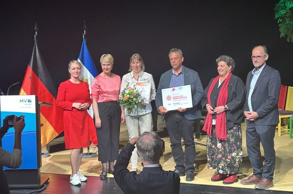 Gruppenfoto auf einer Bühne: Sieben Personen stehen nebeneinander vor dunklem Hintergrund. Links steht ein Rednerpult mit dem Logo „MV tut gut.“ Daneben sind zwei Fahnen zu sehen, eine in Schwarz‑Rot‑Gold und eine blau‑weiße. In der Mitte hält eine Person einen Blumenstrauß, eine weitere hält eine Urkunde, eine andere ein gerahmtes Schild mit Text und Logos. Die Personen tragen unterschiedliche Kleidung in Rot‑, Grau‑, Blau‑ und Schwarztönen. Im Vordergrund ist von hinten eine Person mit erhobener Kamera zu sehen, die das Foto aufnimmt. Rechts im Hintergrund stehen mehrere bunte Stühle.