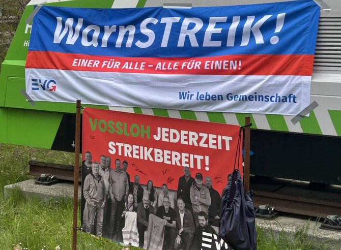 An einem grünen Schienenfahrzeug hängt ein großes Banner mit der Aufschrift „WarnSTREIK! Einer für alle – alle für einen! Wir leben Gemeinschaft“ und dem EVG‑Logo. Davor steht ein rotes Plakat mit der Aufschrift „VOSSLOH JEDERZEIT STREIKBEREIT!“ sowie mehreren abgebildeten Personen und Unterschriften. Im Vordergrund sind Gleise und Gras zu sehen.