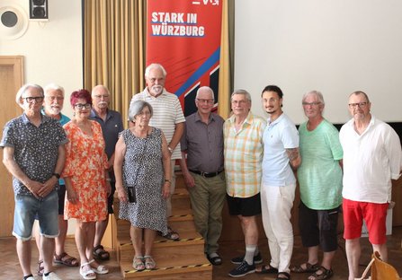 Eine Gruppe von Menschen steht nebeneinander vor einem roten Banner mit der Aufschrift „STARK IN WÜRZBURG“ und blickt in die Kamera. Sie tragen unterschiedliche Kleidung, darunter Hemden, Kleider und Shorts.