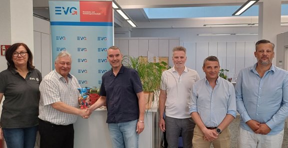 Sechs Personen stehen in einem hellen Büro vor einem Roll-up mit mehrfach sichtbarem „EVG“-Logo. Zwei Personen in der Mitte schütteln sich die Hand und halten gemeinsam eine kleine bunte Figur. Im Hintergrund sind Pflanzen, Schränke und Deckenleuchten zu sehen.