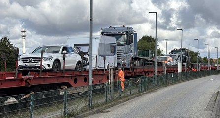Beladener DB-Flachwagen mit Pkw, Wohnwagen, Lkw und weiteren Fahrzeugen. Eine Person in orangefarbener Warnkleidung steht neben dem Zug. Entlang des Gleises verläuft ein Zaun, daneben eine Straße mit Laternenmasten unter bewölktem Himmel.