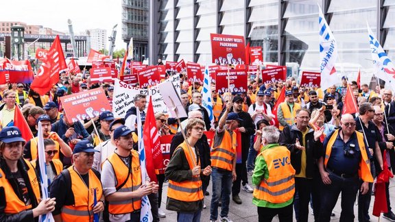 Große Menschenmenge auf einem Platz vor modernen Gebäuden. Viele Personen tragen orangefarbene Warnwesten und dunkle Kappen. Zahlreiche rote, weiße und blaue Fahnen sowie rote Schilder werden hochgehalten. Auf mehreren Schildern ist der Text „JEDEN KAMPF WERT“ sowie Ortsnamen wie „PADERBORN“, „MÜNSTER“ und „WOLFSBURG“ sichtbar. Im Hintergrund ist ein Gebäude mit der Aufschrift „POTSDAMER PLATZ“ zu sehen.