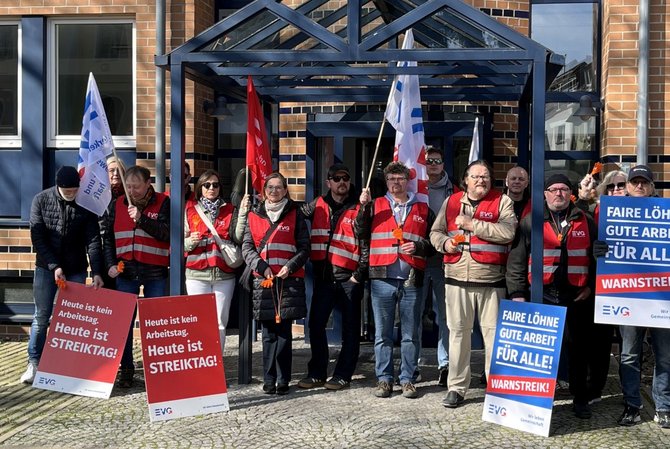 Gruppe von etwa zwölf Personen steht dicht nebeneinander vor dem Eingang eines Backsteingebäudes mit Glasvordach. Die Personen tragen rote Warnwesten mit EVG‑Logo und halten Fahnen sowie Schilder. Mehrere Schilder zeigen gut lesbaren Text: „Heute ist kein Arbeitstag. Heute ist STREIKTAG!“ sowie „FAIRE LÖHNE GUTE ARBEIT FÜR ALLE! WARNSTREIK!“. Die Fahnen sind überwiegend weiß oder blau mit EVG‑Beschriftung. Der Boden besteht aus Pflastersteinen; hinter der Gruppe sind eine Glastür und Fenster des Gebäudes sichtbar.