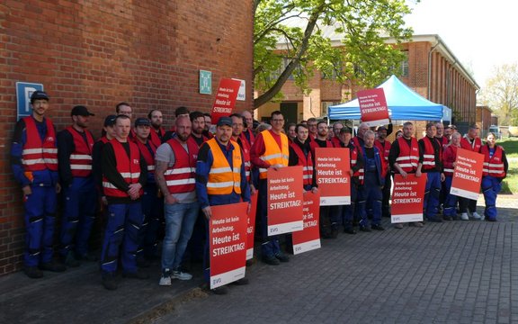 Gruppe von Personen in roter Warnweste und blauer Arbeitskleidung steht dicht nebeneinander vor einer roten Backsteinwand im Außenbereich. Mehrere Personen halten rechteckige rote Schilder mit weißer Aufschrift „Heute ist kein Arbeitstag. Heute ist STREIKTAG!“ und kleinem „EVG“-Logo am unteren Rand. Im Hintergrund sind ein Gebäude aus rotem Backstein, ein Baum mit grünem Laub und ein blaues Pavillonzelt zu sehen.
