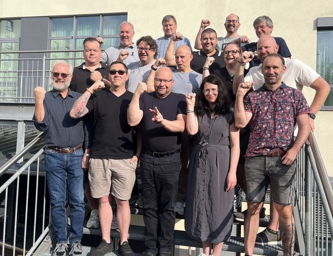 Gruppe von 14 Personen steht auf einer Metalltreppe vor einem Gebäude mit großen Fenstern. Die Personen sind in mehreren Reihen angeordnet und halten jeweils einen angewinkelten Arm mit geballter Faust nach oben. Einige tragen Brillen. Die Kleidung ist überwiegend dunkel oder grau, einzelne Oberteile sind gemustert. Helles Sonnenlicht fällt von der rechten Bildseite auf die Gruppe und erzeugt deutliche Schatten. Geländer und Gebäudefassade bilden den Hintergrund.