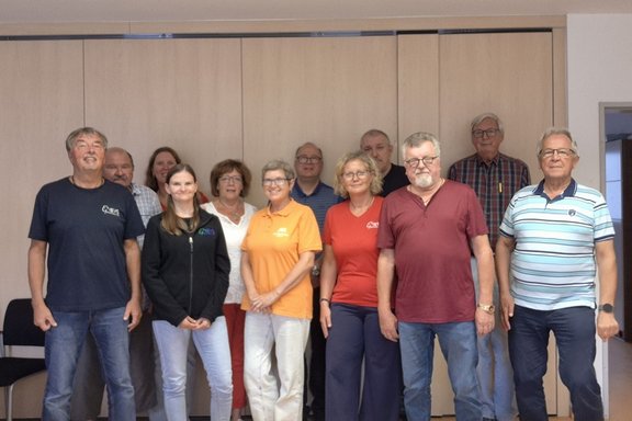 Elf Personen stehen in zwei Reihen in einem Besprechungsraum vor einer geschlossenen, hellen Schrankwand und blicken in die Kamera. Die Gruppe besteht aus elf Personen, die für ein gemeinsames Gruppenfoto aufgestellt sind. Sechs Personen stehen in der vorderen Reihe, fünf Personen dahinter. Die Personen tragen überwiegend Freizeitkleidung in verschiedenen Farben, darunter Dunkelblau, Schwarz, Orange, Rot, Bordeauxrot und gestreifte Oberteile.