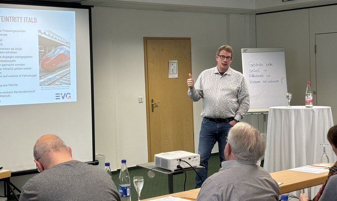 Seminarraum mit Leinwand und Präsentation. Eine Person steht vor der Leinwand, zeigt mit einer Hand nach hinten und spricht zu mehreren Teilnehmenden, die an Tischen sitzen. Auf der Leinwand ist eine Folie mit einem Zugbild und dem EVG-Logo zu sehen. Rechts steht ein Flipchart mit handschriftlichem Text: „Getränke bitte selbst im Kühlschrank im Vorraum entnehmen!“. Im Vordergrund stehen ein Beamer auf einem kleinen Tisch, Wasserflaschen, Gläser und Unterlagen.