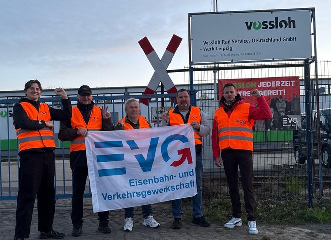 Mehrere Personen stehen vor einem Metallzaun und tragen orange Warnwesten. Einige halten eine weiße Fahne mit der Aufschrift „EVG Eisenbahn- und Verkehrsgewerkschaft“. Hinter dem Zaun ein Schild: „Vossloh Rail Services Deutschland GmbH – Werk Leipzig“. Daneben ein weiteres Schild mit der Aufschrift „Vossloh jederzeit einsatzbereit!“. Im Hintergrund industrielle Anlagen, ein Andreaskreuz und ein bewölkter Himmel.