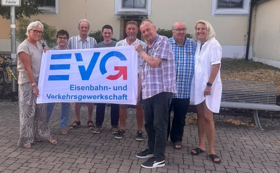 Acht Personen stehen auf einem gepflasterten Platz vor einer Hausfassade und halten gemeinsam ein weißes Banner. Auf dem Banner steht in großen blauen und roten Buchstaben „EVG“ sowie darunter „Eisenbahn- und Verkehrsgewerkschaft“. Links ist ein Straßenschildmast teilweise sichtbar, rechts eine Metallbank. Im Hintergrund sind Fenster, Sträucher und die Gebäudefassade zu sehen.Geben Sie Ihr Feedback zu BizChat ab.