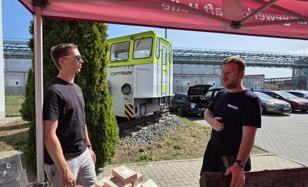 Zwei Personen stehen unter einem roten Pavillon und unterhalten sich. Im Vordergrund befinden sich mehrere Schachteln auf einem Tisch. Im Hintergrund sind die grün-weiße Lokomotive mit der Aufschrift „CAPTRAIN“, geparkte Fahrzeuge, Industrieanlagen und Gebäude zu sehen.