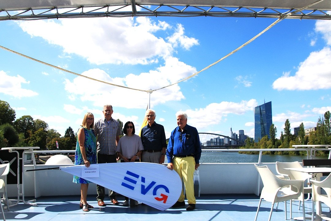 Vier Personen stehen auf dem Außendeck eines Schiffes hinter einer EVG-Fahne und posieren für ein Gruppenfoto vor der Frankfurter Skyline. Die Gruppe befindet sich auf einem hellblauen Schiffsdeck. Vor den Personen liegt eine weiße Fahne mit dem blau-roten EVG-Logo. An der Fahne ist ein Schild befestigt, auf dem „EVG Frankfurt“ und „60-jähriges Jubiläum“ zu lesen ist. Hinter der Gruppe steht ein Mast mit einer Deutschlandflagge.
