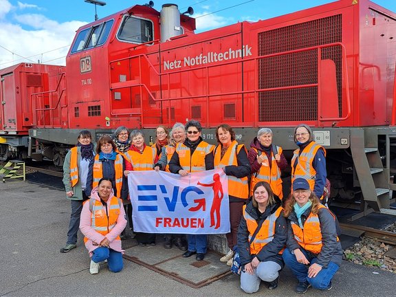 Eine Gruppe von weiblichen Beschäftigten in orangefarbenen Warnwesten steht vor einem roten Eisenbahnfahrzeug und hält ein Banner mit der Aufschrift „EVG Frauen“. Die Personen stehen und knien gemeinsam auf einem Bahngelände. Hinter ihnen ist ein großes rotes Schienenfahrzeug mit der Aufschrift „Netz Notfalltechnik“ zu sehen. Das Banner ist weiß, mit blau‑roter Schrift und einer stilisierten Figur. Einige Personen tragen zusätzlich Jacken und Schals. Im Hintergrund sind Gleise und weitere Eisenbahntechnik erkennbar.
