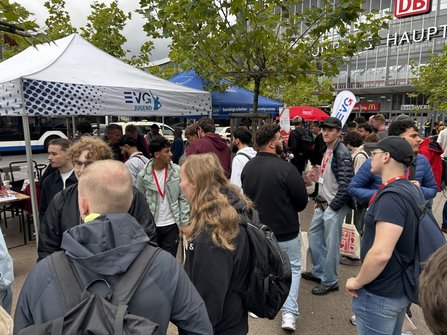 Menschenmenge auf einem Platz mit Bäumen vor einem großen Gebäude mit Glasfassade. Im Vordergrund und um einen weißen Pavillon mit der Aufschrift „EVG JUGEND“ stehen zahlreiche Personen mit Namensschildern an roten Bändern, einige tragen helle Stofftaschen. Dahinter sind ein blauer Pavillon, eine weiße Fahne mit EVG-Logo und weitere Informationsstände zu sehen. Im Hintergrund befindet sich ein Gebäude mit einem roten „DB“-Logo und teilweise sichtbarer Beschriftung „HAUPT…“. Der Himmel ist stark bewölkt.