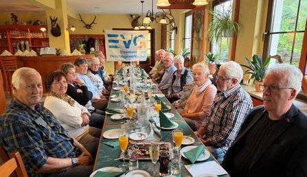 Mehrere Personen sitzen an einem langen, gedeckten Tisch in einem Gastraum. Auf dem Tisch stehen Gläser, Flaschen, Kerzen, Teller und gefaltete grüne Servietten. An der Stirnseite des Raums hängt ein Banner mit der Aufschrift „EVG Eisenbahn- und Verkehrsgewerkschaft“. An den Wänden befinden sich Holzregale, Garderobenhaken, Geweihe und weitere Dekorationen. Große Fenster verlaufen entlang einer Seite des Raums.