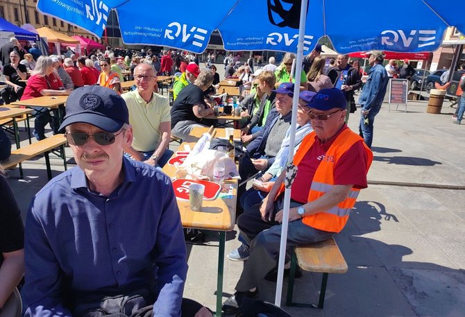 Außenaufnahme eines Platzes mit vielen Menschen an Biertischgarnituren. Mehrere Personen sitzen an Holztischen unter blauen Sonnenschirmen mit der Aufschrift „RWE“. Auf den Tischen stehen Getränke, Becher und Taschen. Einige Personen tragen orangefarbene Warnwesten mit der Aufschrift „EVG“. Im Hintergrund sind weitere Stände, Menschen und Gebäude sichtbar.