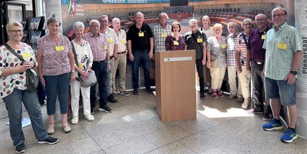 Gruppenfoto mit etwa 15 Personen in einer Eingangshalle. Die Gruppe steht um ein Rednerpult vor einer Wandgrafik mit dem Schriftzug „Landtag Nordrhein-Westfalen“ und einer Darstellung eines Plenarsaals. Die Personen tragen gelbe Namensschilder.