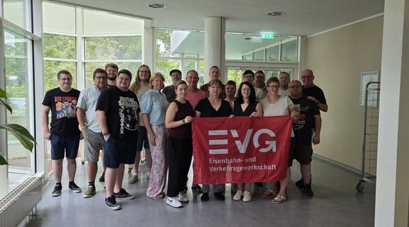 Gruppe von etwa 18 Personen in einem hellen Innenraum mit großen Fenstern. In der Mitte halten mehrere Personen eine rote Fahne mit der Aufschrift „EVG Eisenbahn- und Verkehrsgewerkschaft“. Links und rechts stehen weitere Personen. Im Hintergrund sind Fenster, eine Glastür, Säulen und Pflanzen zu sehen.