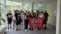 Gruppe von etwa 18 Personen in einem hellen Innenraum mit großen Fenstern. In der Mitte halten mehrere Personen eine rote Fahne mit der Aufschrift „EVG Eisenbahn- und Verkehrsgewerkschaft“. Links und rechts stehen weitere Personen. Im Hintergrund sind Fenster, eine Glastür, Säulen und Pflanzen zu sehen.