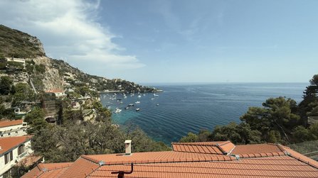 Blick von erhöhter Position über eine Küstenbucht mit ruhigem Meer. Im Vordergrund sind rote Ziegeldächer zu sehen, links ein bewaldeter Hang mit Gebäuden. In der Bucht liegen zahlreiche Boote vor der Küste. Der Himmel ist überwiegend klar mit einigen Wolken.