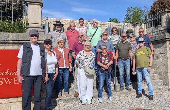 Gruppe von etwa zwanzig Personen, die in mehreren Reihen auf einer steinernen Treppe im Außenbereich stehen. Hinter der Gruppe befindet sich ein schmiedeeisernes Tor und eine helle Steinmauer. Links im Vordergrund ist ein rotes Schild mit weißer, nur teilweise sichtbarer Schrift angebracht. Die Personen tragen unterschiedliche Freizeitkleidung wie Hosen, Hemden, Jacken und T‑Shirts. Der Boden besteht aus hellem Steinpflaster, darüber ist ein blauer Himmel sichtbar.