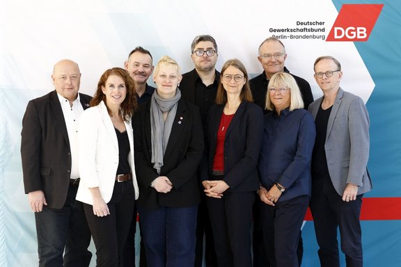 Gruppenfoto mit elf Personen, die in zwei Reihen vor einem hellen Hintergrund stehen. Im Vordergrund stehen sechs Personen nebeneinander, im Hintergrund fünf weitere. Alle tragen überwiegend dunkle, formelle Kleidung wie Blazer, Jacken und Hemden; einzelne Kleidungsstücke sind hell oder farbig, darunter ein weißer Blazer, ein rotes Oberteil und ein grauer Schal. Mehrere Personen haben die Hände vor dem Körper gefaltet. Im oberen rechten Bereich des Hintergrunds sind das Logo „DGB“ sowie der Text „Deutscher Gewerkschaftsbund Berlin-Brandenburg“ zu sehen. Der Hintergrund ist weiß mit dezenten geometrischen Farbflächen in Blau- und Rottönen.