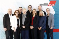 Gruppenfoto mit elf Personen, die in zwei Reihen vor einem hellen Hintergrund stehen. Im Vordergrund stehen sechs Personen nebeneinander, im Hintergrund fünf weitere. Alle tragen überwiegend dunkle, formelle Kleidung wie Blazer, Jacken und Hemden; einzelne Kleidungsstücke sind hell oder farbig, darunter ein weißer Blazer, ein rotes Oberteil und ein grauer Schal. Mehrere Personen haben die Hände vor dem Körper gefaltet. Im oberen rechten Bereich des Hintergrunds sind das Logo „DGB“ sowie der Text „Deutscher Gewerkschaftsbund Berlin-Brandenburg“ zu sehen. Der Hintergrund ist weiß mit dezenten geometrischen Farbflächen in Blau- und Rottönen.