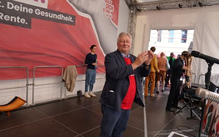 Person auf einer Bühne in dunklem Anzug und rotem T-Shirt mit erhobenen Händen beim Klatschen. Im Hintergrund befindet sich ein großes rotes Banner mit teilweise sichtbarem Text „Deine Gesundheit. Rente.“ sowie „MACH DICH STARK MIT UNS“. Rechts sind Mikrofone, ein Blechblasinstrument auf einem Ständer und mehrere weitere Personen zu sehen. Die Bühne ist von einer Metallkonstruktion und weißen Zeltwänden umgeben.