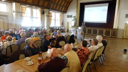 Großer Saal mit Bühne, rotem Vorhang und heruntergelassener Projektionsleinwand. Eine Person steht vor der Bühne und spricht zu den an Tischen sitzenden Personen. Zahlreiche Getränke, Tassen und Tischaufsteller befinden sich auf den Tischen. Hohe Fenster an der linken Wand sorgen für helle Beleuchtung im Raum.