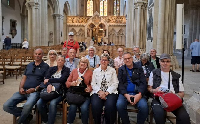 Gruppe von etwa fünfzehn Personen, die in mehreren Reihen auf Holzbänken im Inneren einer großen Kirche sitzen. Der Raum ist von hohen Steinpfeilern, Rundbögen und einer reich verzierten Altarwand geprägt. Im Hintergrund sind bunte Glasfenster zu sehen, durch die Licht in den Raum fällt. Die Personen tragen unterschiedliche Freizeitkleidung und halten teils Taschen oder Gegenstände in den Händen. Weitere leere Holzbänke und einzelne stehende Personen sind im Hintergrund sichtbar.
