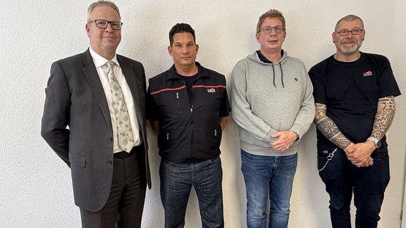 Vier Personen stehen nebeneinander vor einer weißen, leicht strukturierten Wand in einem Innenraum. Im Vordergrund sind alle vier Personen vollständig von Kopf bis etwa zu den Oberschenkeln sichtbar, frontal zur Kamera ausgerichtet, mit den Händen vor dem Körper oder hinter dem Rücken.