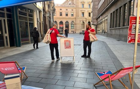 In einer gepflasterten Einkaufspassage stehen zwei Personen in roten T‑Shirts mit hellen Stofftaschen. Zwischen ihnen befindet sich ein Aufsteller mit einem Plakat und QR‑Code. Im Vordergrund stehen zwei rote Liegestühle. Im Hintergrund sind Gebäude, Schaufenster und einzelne vorbeigehende Personen zu sehen.