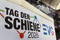 Ein Schild zum „Tag der Schiene 2026“ ist neben dem Logo der Eisenbahn- und Verkehrsgewerkschaft (EVG) auf einem Informationsstand zu sehen. Das Bild zeigt einen nahen Ausschnitt eines Standes oder einer Präsentationsfläche. Im Vordergrund befindet sich ein großes Schild mit der Aufschrift „TAG DER SCHIENE 2026“. Daneben ist das blau-rote Logo der EVG zu sehen. Beide Elemente nehmen den größten Teil des Bildes ein.