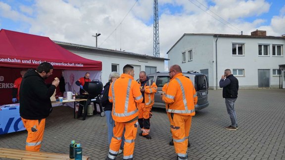 Mehrere Personen in orangefarbener Warnkleidung stehen auf einem gepflasterten Hof und unterhalten sich. Links befindet sich ein rotes Zelt mit der Aufschrift „Eisenbahn- und Verkehrsgewerkschaft Halle“, darunter ein Tisch mit Gegenständen und ein Grill. Im Hintergrund sind ein grauer Transporter, weitere Personen in dunkler Kleidung, ein Strommast sowie flache Gebäude mit Fenstern zu sehen. Der Himmel ist teilweise bewölkt.