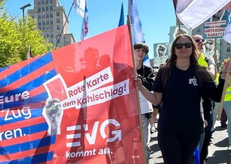 Nahaufnahme eines roten Banners, das von mehreren Personen getragen wird. Auf dem Banner steht „Rote Karte dem Kahlschlag!“ sowie „EVG kommt an.“ Daneben sind weitere Fahnen und Plakate zu sehen. Im Hintergrund befinden sich weitere Teilnehmende eines Demonstrationszuges und städtische Gebäude.