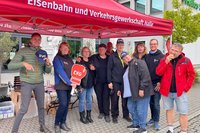 Eine Gruppe von Personen steht unter einem roten Pavillon mit der Aufschrift „Eisenbahn und Verkehrsgewerkschaft Halle“. Einige Personen halten Schilder oder Karten mit dem Logo „EVG“. Im Hintergrund stehen Tische mit Materialien, Kartons sowie ein Gebäude mit Fenstern.