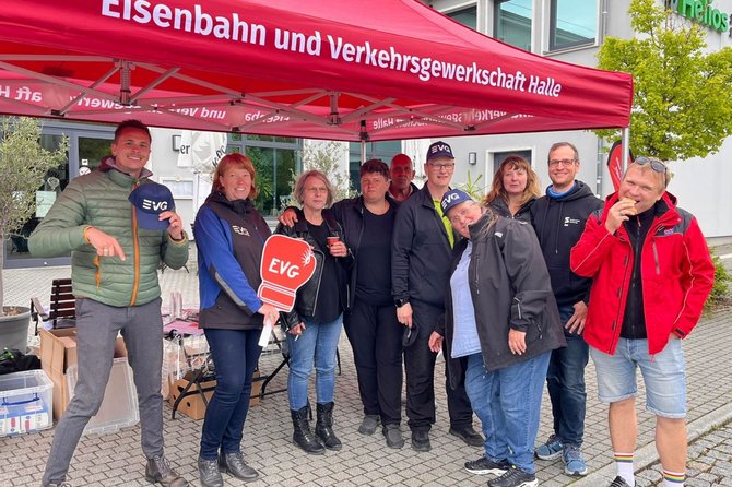 Eine Gruppe von Personen steht unter einem roten Pavillon mit der Aufschrift „Eisenbahn und Verkehrsgewerkschaft Halle“. Einige Personen halten Schilder oder Karten mit dem Logo „EVG“. Im Hintergrund stehen Tische mit Materialien, Kartons sowie ein Gebäude mit Fenstern.