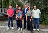 echs Personen stehen im Freien auf einem gepflasterten Weg vor dichtem Grün mit Bäumen und Sträuchern. Links im Hintergrund befindet sich eine Metallbrücke über einen schmalen Wasserlauf. Die Personen tragen unterschiedlich farbige Kleidung, darunter ein auffällig gemustertes Oberteil in der Bildmitte, zwei dunkle Sakkos sowie mehrere Freizeitoberteile. Im Hintergrund sind zwischen den Bäumen Teile eines Gebäudes sichtbar. Tageslicht, Gruppenaufnahme.