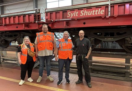 Vier Personen in orangefarbenen Warnwesten stehen vor einem roten Eisenbahnwagen mit der Aufschrift „Sylt Shuttle“. Die Aufnahme entstand in einer Werkhalle mit Gleisanlagen, Industrielampen und großen Fenstern im Hintergrund.