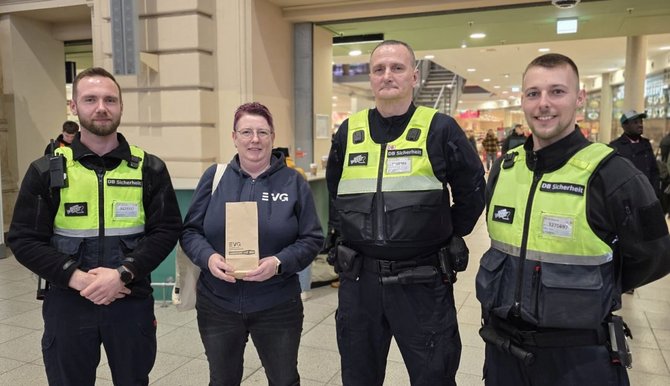 Eine Person in dunkelblauer Kleidung hält eine kleine Papiertüte mit einem EVG‑Logo und steht in einem Bahnhofsgebäude. Links und rechts stehen mehrere Personen in DB‑Sicherheit‑Uniformen mit Warnwesten und Ausrüstung. Im Hintergrund sind Geschäfte, Beleuchtung und ein offener Bereich des Bahnhofs zu sehen.