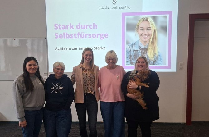 Fünf Personen stehen nebeneinander vor einer Projektionsfläche in einem Seminarraum. Auf der Folie ist der Titel „Stark durch Selbstfürsorge“ und der Untertitel „Achtsam zur inneren Stärke“ zu lesen; oben befindet sich ein Logo mit dem Text „Julia Jeschke Life‑Coaching“. Rechts auf der Folie ist ein gerahmtes Porträt abgebildet. Eine der Personen hält einen kleinen Hund im Arm.